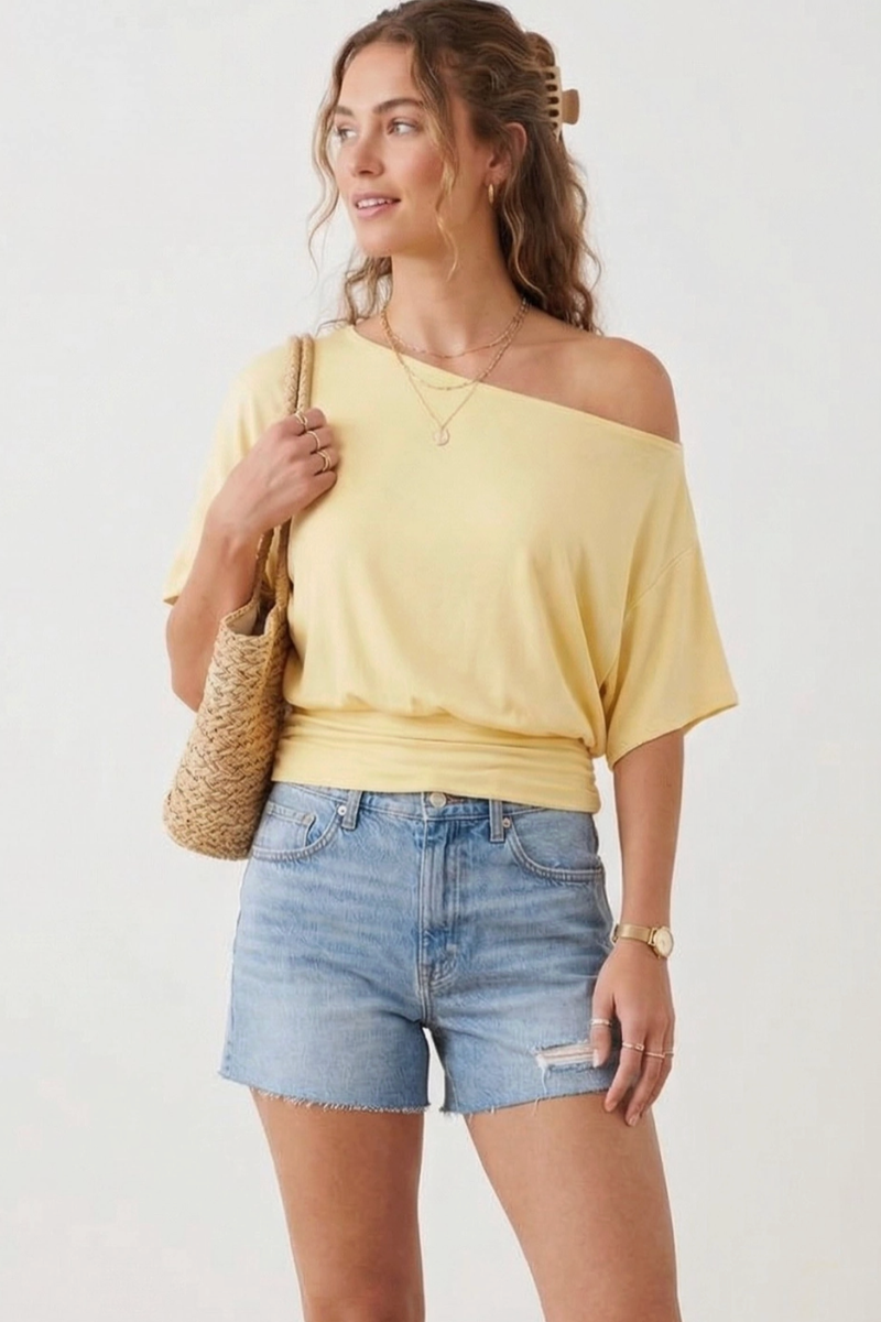 Woman wearing a yellow off-shoulder top and denim shorts, holding a woven bag.